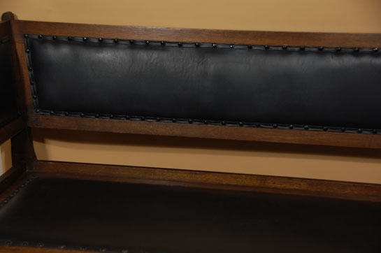 This settle was made from quarter-sawn oak. The arms are splayed outward in a fashion that provides for more comfort. One does not feel being boxed in when sitting near an arm. The back panel, too, is splayed outward for added comfort. The seat, back and interior arm panels are upholstered in black leather or faux leather. The settle does not display a manufacturer’s label. It is a period piece, dating the early part of the 20th century.