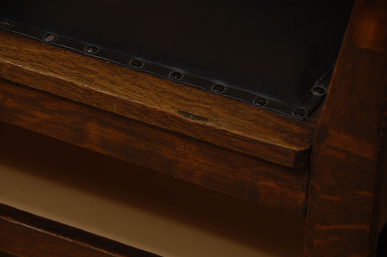 This settle was made from quarter-sawn oak. The arms are splayed outward in a fashion that provides for more comfort. One does not feel being boxed in when sitting near an arm. The back panel, too, is splayed outward for added comfort. The seat, back and interior arm panels are upholstered in black leather or faux leather. The settle does not display a manufacturer’s label. It is a period piece, dating the early part of the 20th century.