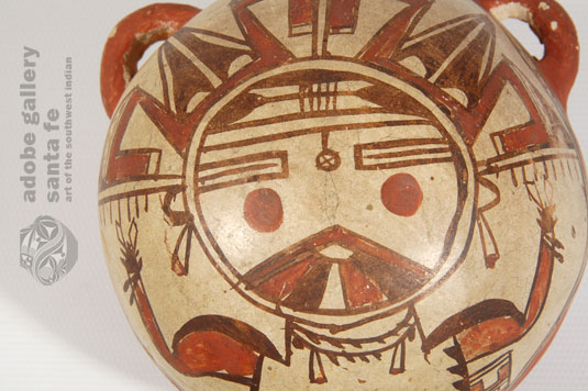 The upper two thirds of the domed body is where Nampeyo displayed her artistic design of a Palhikmana Katsina in almost full perspective.  Her face is at the center of the dome and an elaborate tableta fills the upper half of the area, with the katsina’s body filling in the lower half. She is provided with a traditional Hopi homespun manta, a double string of turquoise nuggets with jaclas as a necklace, and jaclas earrings.  The katsina’s chin displays the traditional rainbow feature of that katsina.