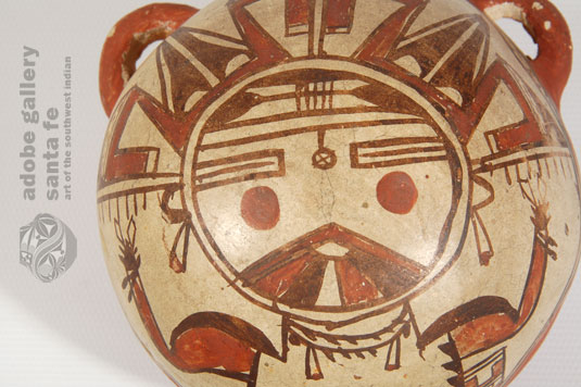 The upper two thirds of the domed body is where Nampeyo displayed her artistic design of a Palhikmana Katsina in almost full perspective.  Her face is at the center of the dome and an elaborate tableta fills the upper half of the area, with the katsina&rsquo;s body filling in the lower half. She is provided with a traditional Hopi homespun manta, a double string of turquoise nuggets with jaclas as a necklace, and jaclas earrings.  The katsina&rsquo;s chin displays the traditional rainbow feature of that katsina.