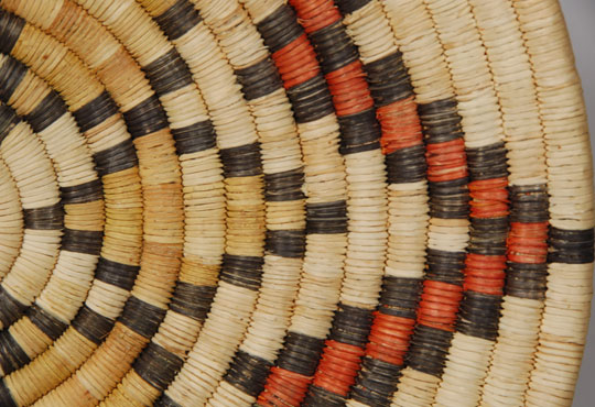 Coiled basketry of this type is made exclusively in Second Mesa villages on the Hopi Reservation. Using an awl, the basket maker pushes the yucca strip into the adjoining stitch of the previous coil. This process is repeated one stitch at a time throughout the entire process of making the basket.  Such an effort is very time consuming and a plaque such as this one probably took the weaver more than a month to complete the weaving process alone, not counting the months of gathering materials, drying the yucca leaves, and then splitting them to a thin, consistent width, dying some of them, and preparing the grass for the foundation.  I have watched a number of women at Second Mesa villages make baskets over the years, and I am in awe of their patience and fortitude.  This plaque features a central design of radiating stars.  The outer star consists of alternating black and red blocks and the inner one of yellowed and dyed yucca.  The basket is somewhat shallow but does have some concavity.  There is a loop woven into the back to facilitate hanging.  Condition:  very good condition considering its age of approximately 35 years.  Provenance: from the collection of the Balcomb family  Recommended Reading:  Hopi Basket Weaving: Artistry in Natural Fibers by Helga Teiwes 