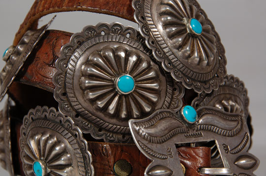 This fine old concha belt is chisel-stamped repoussé and filed.  Each of the 10 conchas features a sunburst at its dome capped with a single blue turquoise cab secured in a silver bezel.  The buckle is also of the same construction with two turquoise cabs.  This style is the epitome of the finest concha belts made in the mid-20th century.   The conchas are not overbearingly large and each is attached to the leather belt with a copper sleeve.    Silver jewelry from the early- to mid-20th century was still being produced by many Navajo at home in their hogans.  There was no widespread production by Natives working for outlets such as The Fred Harvey Company using company-supplied materials.  Belts such as this one were most certainly made by an individual silversmith and possibly sold to a trading post or dealer or an individual collector.  The early interest by tourists was a stimulus to fine craftsmanship.  It was only in the 1970s that quality gave way to quantity.  Condition:  the belt is in excellent condition  Provenance: from the collection of the Balcomb family  Recommended Reading: Indian Silver Jewelry of the Southwest 1868-1930 by Larry Frank Concho or Concha?  You will hear both words (we say conca), but they refer to the same thing.  Some history:  The word concho comes from the Spanish "concha" which actually means "conch" or "seashell" but has come to mean round, domed, or oval disks of silver used to decorate horse saddles or bridles and even on clothing.  It’s also seen as jewelry such as pendants, pins and even bolo ties.  What is most striking is when it’s seen adorning leather belts.  Perhaps you will even hear a single “concha” referred to as concha instead of concho. Concho belts are a long-time Navajo tradition yet it has been suggested (unknown source) that the Navajo borrowed conchos from Mexican tack items or from the Plains Indians.  The earliest conchos were silver dollars (coin silver) that were hammered, then stamped and edged, then slotted and strung together on a piece of leather.  Eventually, copper loops were added to the back of the conchos so that the conchos could be slipped onto a leather belt. Features of a Concho Belt: Concho belts can be a continuous row of conchos or could have spacers in between the conchos. The spacers can of various shapes but traditionally are butterflies and it is easy to see why they are called that when you look at the shape of them. The conchos and the butterflies are sometimes backed by leather which highlights the silver work and also protects the edges of the silver from bumping, wear or bending. Can a man or a woman wear a concho belt?   Concho belts are unisex and can be worn with jeans as well as dresses.  It is very “Santa Fe” to add a concho belt to your attire and can even be included with any clothing that is black in color.  When this is done, this creates what is considered “Santa Fe Formal” wear and is usually very stunning.  This approach really draws attention to the wearer’s concho belt and oftentimes, great conversation.   