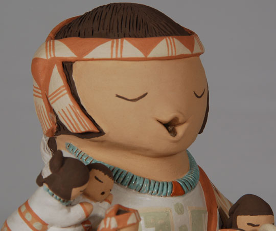 Stella Teller has a long career of making storyteller figurines and has consistently won prizes for them since she made the first one in 1978, the same year Adobe Gallery opened its doors in Albuquerque.  Her great-grandmother, Marcellina Jojola, her grandmother, Emeklia Lente Carpio, and her mother, Felicita Jojola, were all potters.  Her pottery is distinguished from traditional Isleta Polychrome wares by its distinctive colors, which she says are all natural.  The light gray, which has become her trademark, is produced by mixing white clay with manganese.  She was one of the first potters to insert turquoise cabs into the clay.  Not to be considered parochial, Teller expanded the repertoire to include storytellers representing Navajo, Isleta Pueblo, Apache and Hopi males and females.  She is credited with making the first storyteller to represent a Navajo.  This storyteller is dressed in the garb of a Navajo man.  He has four boys and two girls clamoring all over him.  The boys have their interest focused on the large drum he holds.  The girl’s minds are elsewhere.  The adult wears a painted headband, a chonga hair style, a blanket draped over his left shoulder, a turquoise necklace and traditional moccasins on his crossed legs.    Condition: this storyteller is new and in original condition  Provenance: from the artist  Recommended Reading:  The Pueblo Storyteller by Barbara Babcock 
