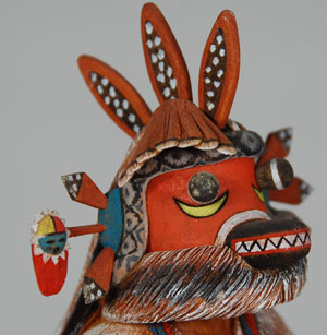 Today, the Scavenger (Uhuhu) Katsina performs as a guard or disciplinarian.  His staff and knife are indications of his control over Katsina dances.  Also, he accompanies the Ogre Woman when she gathers all the items given to her during a ceremony.  After she has filled her basket, the Scavenger Katsina picks up the remaining items and fills his basket.  In this capacity, he is considered a part of the Ogre family of Katsinas.  Historically, the Scavenger was considered a looter or scavenger of the Awatowi ruins, picking up anything of interest.  This is an all-wood carving by Brian Laban completed in 1997.  The basic carving is from a single piece of cottonwood root, with the staff, knife and protrusions on the mask being added items.  The detail achieved by the carver is absolutely extraordinary.  He even carved an arm band on the right arm of the katsina even though it is completely covered by the sheep skin cape.  It is details such as this that have made Laban one of the finest carvers on the reservation.  The face of the katsina features a long snout with teeth, two bug eyes under which are new moons, feathers on both sides as well as the top of the head, and a sun symbol jutting out from one side.  Long black hair drapes down the back.  A serpent is draped over the forehead.  On the Scavenger’s back hangs a basket held in place by the red woven sash.  The cape over the shoulders represents the sheep skin cape worn by the katsina.  A coral and turquoise necklace adorns the neck and shell bracelets are on both wrists.  A beautifully carved Hopi embroidered sash hangs in a flowing manner on the right side of the katsina and a concha belt wraps around the waist of the doll.  On the left side of the waist is a shield with a star and moon.  A foxtail hangs down the back side and traditional moccasins are on the feet.  There is absolutely nothing omitted on this carving.  Laban has done a marvelous job.  Condition: original condition  Provenance: Adobe Gallery purchased this carving from the artist in 1997 and sold it to a New York client who now has brought it back to us for re-sale. Recommended Reading:  Hopi Katsina: 1,600 Artist Biographies by Gregory and Angie Schaaf 