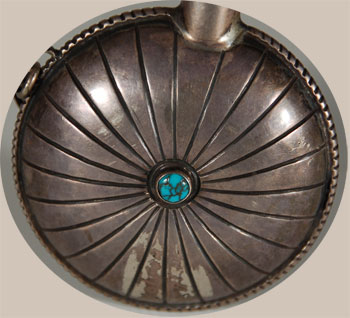 Silversmithing among the Navajo began sometime in the mid-1800s, possibly during the time of their internment at Bosque Redondo in southern New Mexico. The early days of silversmithing consisted of jewelry items such as bracelets, necklaces and earrings, and the source of silver was most often Mexican silver coins.  By the beginning of the 20th century, ingot silver and then sheet silver became the source of raw materials, and the demands of the buying public became the inspiration for ideas. In addition to making jewelry, the Navajo expanded into more elaborated items such as horse bridles and ketohs (arm bands) for bow hunting. Eventually, they ventured into making flatware, dishes, serving wares, and such things as silver boxes and desk sets.  One of the things primarily made for their own use were tobacco canteens or flasks. They were generally made by attaching two domed stamped silver concha-shaped discs together and attaching a lidded spout, as was this one. Sometimes, turquoise pieces were added.   This flask has a small beautiful blue turquoise cabochon at the center on front and back. The stamping consists of radial lines from the center. A handmade silver chain secures the canteen stopper.  The weight of this flask would indicate that it was made from ingot silver.  Condition: very good condition Provenance:  from the collection of a Santa Fe family Recommended Reading:  Masterworks and Eccentricities: The Druckman Collection; Navajo and Pueblo Jewelry and Metalwork 1880-1950 by Robert Bauver 
