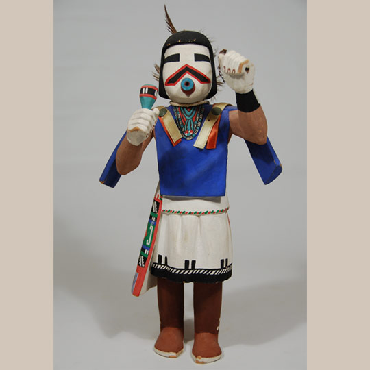 Peter Shelton, Jr. is widely recognized as one of the major carvers of Katsina dolls in the mid-20th century.  He and his brother, Henry Shelton, are names recognized by collectors of older carvings.  He contributed to a style of more detail such as folds in kilts and shirts.  This katsina doll appears to be of the style of his work in the 1950s and 1960s.   When we first received this carving, there was a question in my mind whether it was the First Mesa Shalako Katsina who wears a velvet ribbon shirt or the Navan Katsina who also wears a velvet ribbon shirt.  Neither seemed quite correct but both seemed to be possibilities.  As I always do when I am unsure about a katsina, I consulted with the expert on Hopi and Zuni Katsinas-Chad Burkhardt.  I have known Chad since he was about 10 years old and he would come to the Albuquerque gallery and stand in front of the 10-foot showcase full of Katsina dolls and proceed to name each of them.  It was remarkable that a person of that age was so knowledgeable and interested in such an esoteric subject.  He said this one is the Wakas'la'laiya (Cow Herder) Katsina.   The Wakas'la'laiya Katsina is the side dancer for Wakas'katsinam (Cow Katsinas) when they come as a group.  He has a small round rattle of the type that this doll is holding and is dressed just like this doll.  The Wakas'katsinam represent animal spirit messengers to the Rain gods. Their songs and dance movements are prayers for rain, and are symbolic of animals' self-sacrifice in supplying food to sustain life." (Secakuku)  This carving was very well made.  The katsina doll stands erect, with one foot slightly in front of the other, as if in the process of taking a step.  It stands alone without support or may be hung from the string around its neck.    The shirt and kilt were carved to show folds, and the ribbons over the shoulder are real, not painted. The painting of the embroidered sash is excellent. The carving is signed Hoyesva under one foot.   Condition: He is in very good condition, with no evidence of damage. Provenance: from the Michael Frost estate Recommended Reading: Hopi Katsina: 1,600 Artist Biographies by Gregory Schaaf