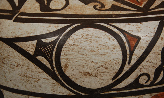 The primary design feature on this jar is what has been named capped spirals and it appears on Zuni pottery dating since 1850.  In this jar, two capped spirals are joined by their long black line which results in a triangular effect.  There are pairs of capped spirals in two rows on the main body separated by a series of orange birds with strong black eyes.  The continuous band of orange birds is broken twice by placement of large medallion figures.  The neck design features the traditional daggers and hachured triangles seen on most Zuni jars of the late 1800s.  The rim of the jar and base are painted black and a puki line is definitely discernible.  The jar certainly dates to no later than 1880.  There have always been strict guidelines at Zuni Pueblo regarding pottery decoration, yet each potter has also always had some degree of freedom. The things that seem to be traditional and inflexible are the manner in which the main body decoration and that of the neck are separated. According to Zuni informants to Ruth Bunzel in 1924 and 1925, it is imperative that the first bit of decoration that goes on any Zuni jar be the black framing line or pair of framing lines that separate the neck from the main body. It is also imperative that this line or pair of lines not be connected. Today we refer to this as a ceremonial break. The informants were explicit in these statements.  Another noticeable aspect on Zuni jars is that there is no correlation between the design of the neck and that of the body of the vessel, except that a certain harmony is preserved between the two. The preferred neck decoration seems to be that shown on this olla. It consists of two designs and they are always used together. One of the designs has a diamond as its center, which represents the bird sling used by boys. The extended parts of this element with the crooks and hachured lines represent the face painting of the Newekwe society. The primary part of the second design around the neck is the triangular arrowhead-looking element that the Zuni women call the thunder knife which the priests use in many ceremonies. Another interesting observation is that the neck of the vessel is generally of the same height as the black underbody of the jar. Traditionally, the body of the vessel is divided into three horizontal bands—two of which are wide and one narrow. Placing medallions at designated spots, as in this jar, frequently breaks up these horizontal bands; however, that further emphasizes the horizontal nature of the design. There are two medallions on this jar and both are simpler in style that medallions of the post-1900 period.  The upper and lower bands on this jar are of unequal width and feature mirror image designs, however, the design in the lower panel is executed in smaller scale due to the shape of the vessel. According to Bunzel, the spiral designs used in the upper and lower bands of a jar will never be used in any other position, such as the neck. Neck designs and body designs are not interchangeable. The spirals here were described to Bunzel as representing the drumstick used by fraternities in their ceremonies in prayers for rain.  A band of red birds, each with black legs and a black dot for an eye, separates the two bands of spirals. The birds are not outlined in black. The use of red elsewhere in the design of the jar is outlined in black, as in the triangles and the medallions.  This jar evidences a puki bulge at the underbody and a prominent ridge at the base of the neck. The base is concave and the rim exhibits a definite lip. It is an excellent example of a 19th century Zuni jar with designs that precede the now-popular deer designs.  Condition: very good condition for a jar of this age.  There is some rim abrasion, probably from use as a water jar and ladle. Reference: The Pueblo Potter: A Study of Creative Imagination in Primitive Art by Ruth Bunzel. Columbia University, New York. Provenance: formerly the property of a family from Gallup, NM who departed the area in mid-20th century. Recommended Reading:  The Pottery of Zuni Pueblo by Lanmon and Harlow 