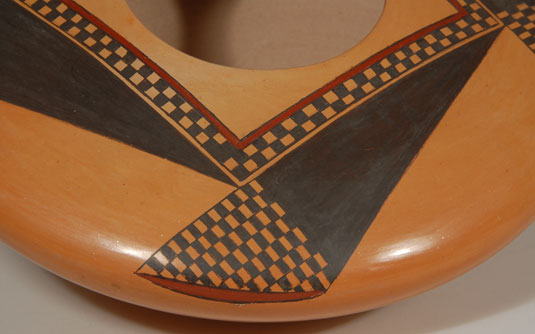 In this seed jar, Dextra adhered to the traditional square enclosing the round opening as seen in other Sikyatki revival designs used by Nampeyo, but then she departed from the Sikyatki designs and created her own.  There is one design repeated four times, once on each leg of the inner square element.  The dark black triangle of each design is capped with rows of black squares in a checkerboard arrangement and then tipped with a small amount of red outlined in black.    The square surrounding the opening is of similar design.  This seed jar is Dextra at her best, simple and elegant.