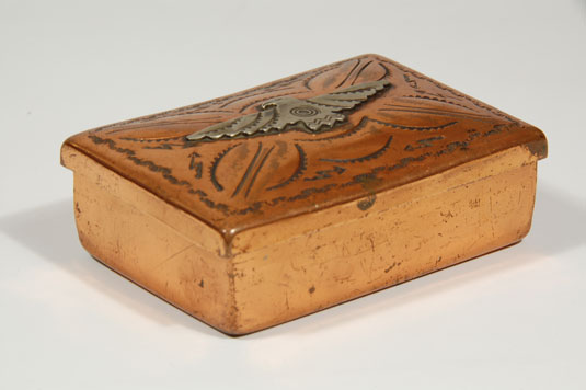 This small box is made of copper with a sterling silver eagle set on the top. Surrounding the eagle are stamped arrows, one of the trademark stamps used by Fred Harvey silversmiths. The top fits snuggly and is connected to the bottom with well-crafted copper hinges. 